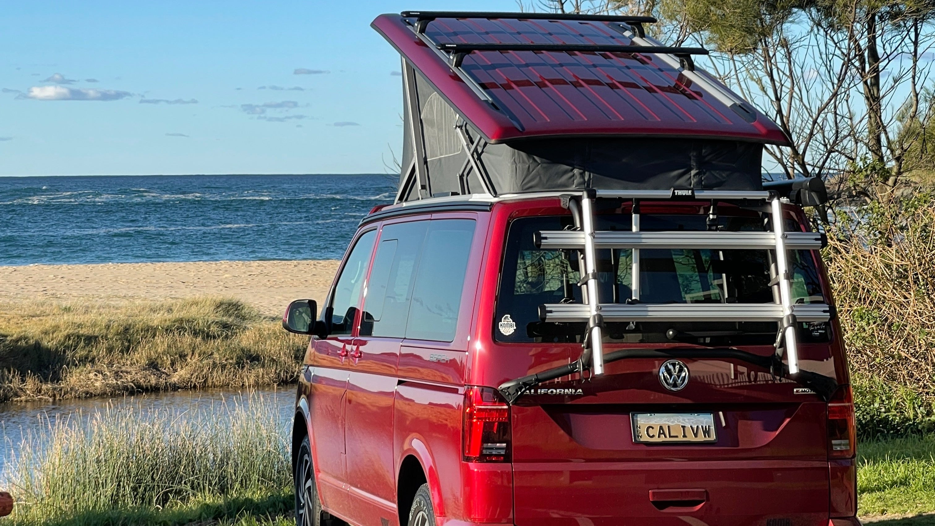 Red VW Kombi Van with a camper shell California parked by the beach with trees in the background powered by K-technik